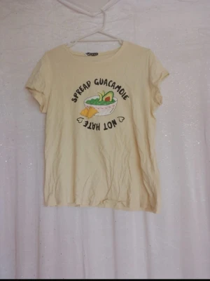 Gul t-shirt med guacamole-tryck - Gul t-shirt med rund hals och softly fit, dekorerad med ett färgg Varje guacamole-try onto och texten 'Spread Guacamole Not Hate' framtill soppar en chill vibe. Perfekt för dig som gillar roliga prints och avslappnad soppar.