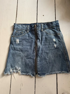 Blå jeanskjol med dragkedja - Trendig blå jeanskjol med rå kant och slitna detaljer. Kjolen har en synlig dragkedja framtill, klassiska fickor och bälteshällor. Perfekt för dig som gillar en avslappnad och edgy stil.