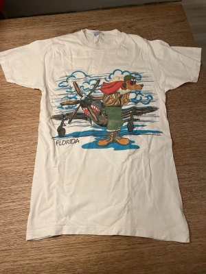 Vit Florida Disney t-shirt med tryck - Säljer en vit t-shirt med coolt färgglatt tryck av ett flygplan med Goofy på framsidan samt texten 'Florida'. T-shirten har klassisk passform och korta ärmar. Perfekt för dig som gillar unika motiv, Disney och retrostil.