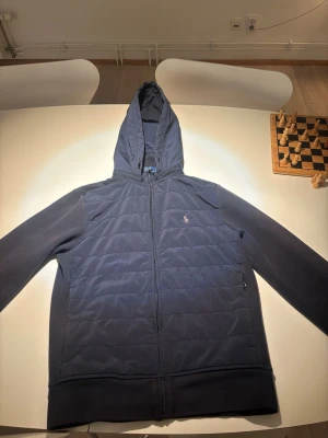 Ralph Lauren  - En Navy Blue Ralph Lauren Cardigan som är i väldigt bra skick, Använts några gånger, inga konstiga lukter, inga hål, ren och fräsch. Pris går att diskuteras               	 •	Materialdesign: Hybridmodell där framsidan är lätt vadderad och quiltad, medan ärmarna och sidorna är i mjukt sweat-/fleecematerial för bättre rörlighet och komfort. 	•	Stängning: Dragkedja fram tills luvans början. 	•	Fickor: Sidofickor med dragkedja. 	•	Logotyp: Grå/vit vanlig Ralph Lauren logga