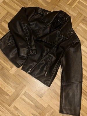  skinnjacka med dragkedjor - Snygg svart/grå skinnjacka i kort modell med bred krage och silvriga dragkedjor på fickorna. Jackan har markerade sömmar och axelklaffar