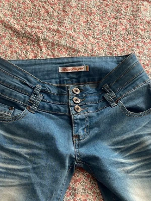 Blå jeans från Row Mayer med låg midja  - Snygga blå jeans från Row Mayer med hög midja och fyra knappar framtill. Jeansen har dekorativa sömmar, bälteshällor och slitningar för en trendig look. Bakfickor med detaljer och klassisk femficksmodell. Perfekta för dig som gillar en modern och cool stil.