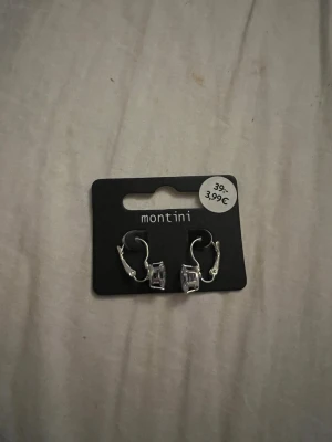 Silvriga örhängen med kubformad sten - Snygga örhängen från Montini i silverfärg med en kubformad, klar sten som ger en stilren och modern look. Örhängena har smidig klickstängning och passar perfekt för dig som gillar minimalistiska accessoarer.