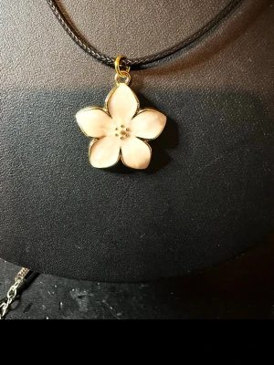 Blommigt halsband med vit blomma - Snyggt halsband med en vit blomma som hänge. Blomman har guldkant och sitter på ett svart band. Perfekt för dig som gillar blommiga detaljer och vill ha något unikt runt halsen.