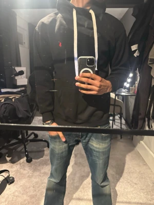 Ralph Lauren hoodie - Säljer en svart hoodie från Polo Ralph Lauren med vit dragsko och klassisk röd logga på bröstet. Hoodien har en stor ficka framtill och ribbade muddar vid ärmslut och nederkant. Perfekt för en chill och stilren look. Storlek M men passar som S