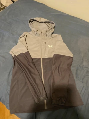 Grå vindjacka Under Armour - Snygg tvåfärgad vindjacka från Under Armour i grått och mörkgrått. Jackan har huva, hel dragkedja framtill och logga på bröstet. Tillverkad i lätt och tåligt material, perfekt för blåsiga dagar. Sportig design med långa ärmar och elastiska muddar.