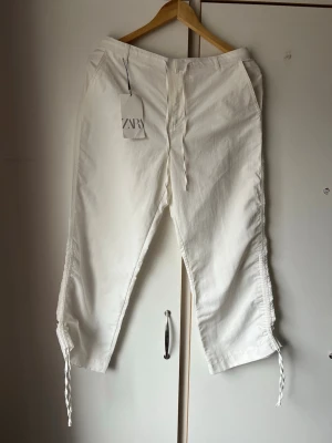 Vit byxa med dragsko från ZARA - Säljer ett par vita byxa från ZARA med dragsko i midjan och längs bensluten. Byxorna har raka ben, fickor på sidorna och är tillverkade i ett mjukt, bekvämt material. Perfekta för en avslappnad och trendig look.