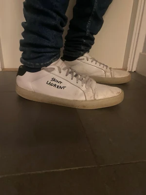 Saint Laurent sneakers - Säljer ett par klassiska vita sneakers från Saint Laurent med svart häl och handskriven logga på sidan. Skorna har lågt skaft, vita snören och en stilren design som passar till det mesta. Tillverkade i läder med gummisula.