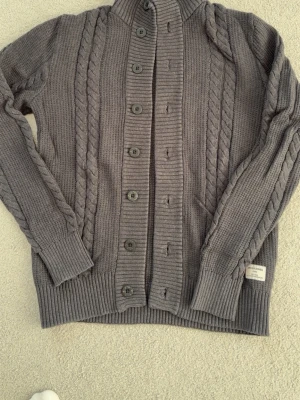 Mörkgrå kofta/ cardigan från Jack&Jones - Mörkgrå kabelstickad kofta från Jack & Jones med stora knappar framtill och ribbade muddar.                 Priset kan diskuteras || Kan även skicka bild på hur de sitter på mig för referens