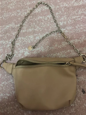 Beige axelremsväska med kedja - Snygg beige axelremsväska i slätt konstläder med silverfärgad kedjerem och dragkedja upptill. Väskan har en minimalistisk design och passar perfekt för dig som vill ha en stilren accessoar till din outfit.