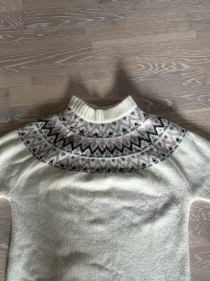 Vit stickad tröja från 157 - Stickad tröja från 157 i vitt med klassiskt nordiskt mönster i svart, grått och beige runt halsen. Tröjan har hög krage och är gjord i ett mjukt, ull-liknande material. Perfekt för kalla dagar och snygg till jeans eller kjol.