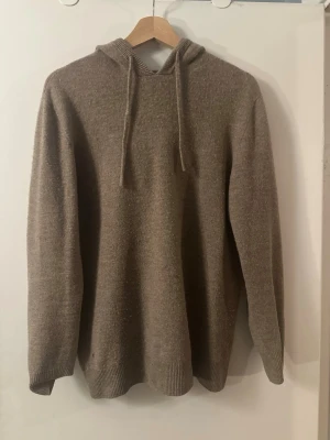 Kashmir beige stickad hoodie med huva - Mysig beige stickad hoodie med huva och dragsko. Tröjan har en avslappnad passform, långa ärmar och ribbade muddar vid ärmslut och nederkant. Perfekt för en chill och stilren look. Materialet känns mjukt och värmande. 50% ull 50% Kashmir 