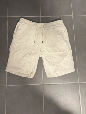 Beige shorts med snörning - Säljer ett par ljusbeige shorts med snörning i midjan och sidofickor. De har en avslappnad passform och är perfekta för varma dagar. Materialet känns lätt och svalt, troligen bomull eller linne. Enkla att matcha till olika outfits!