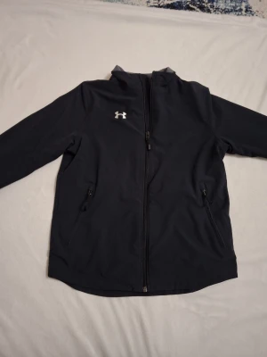 Svart vindjacka från Under Armour - Snygg svart vindjacka från Under Armour med dragkedja framtill och två fickor med dragkedja. Jackan har hög krage och diskret logga på bröstet. Perfekt för dig som vill ha en sportig och stilren look. Jag säljer jackan då den är för liten. Storleken är YLG.