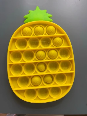 Gul ananas pop it leksak - Rolig pop it leksak i form av en ananas. Gjord av mjuk silikon i gult och grönt, perfekt för att stressa ner eller bara ha kul. Lätt att ta med sig och enkel att använda, tryck på bubblorna för ett tillfredsställande poppande ljud.