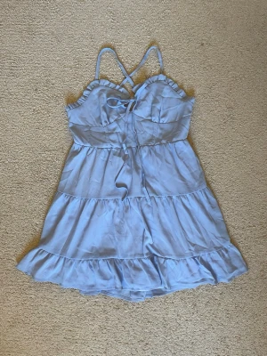 Ljusblå volang gräddig klänning - Söt ljusblå kortklänning med tunna axelband och volangdetaljer. Klänningen har omlottkänsla, knytning fram och öppen rygg med korsade band. Perfekt för sommaren. Materialet känns lätt och luftigt, troligen polyester eller bomull. Använt 2 gånger!
