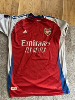 Arsenal matchtröja Adidas röd/blå - Snygg Arsenal matchtröja från Adidas i rött med vita och blå detaljer. Tröjan har klubbmärke på bröstet, Adidas-logga och sponsortryck 'Emirates Fly snapped'. Tillverkad i lätt och ventilerande material, perfekt för fotboll eller träning.