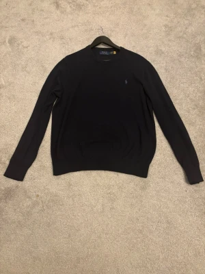 Mörkblå stickad tröja från Polo Ralph Lauren, storlek M - Klassisk mörkblå stickad tröja från Polo Ralph Lauren med rund halsringning och diskret broderad logga på bröstet. Tröjan har långa ärmar och ribbade muddar vid ärmslut och nederkant. Perfekt för lager på lager under kyliga dagar.