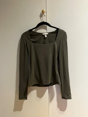 Olivgrön långärmad topp med vid urringning - Snygg olivgrön långärmad topp med bred, fyrkantig urringning. Tillverkad i mjukt och stretchigt material som sitter skönt på kroppen. Perfekt att styla med jeans eller kjol för en trendig look.