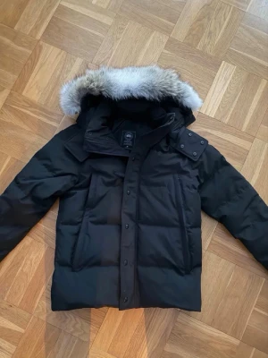 Canada Goose Wyndham (Black Label) - Snygg svart dunjacka från Canada Goose med stor, fluffig huva som har en pälskant. Jackan har två stora framfickor, dragkedja och knappar framtill samt en klassisk boxig passform. Perfekt för kalla vinterdagar. Storlek XL men sitter som storlek L