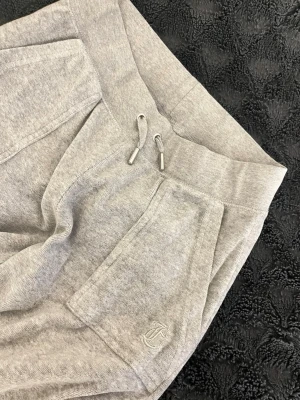 Grå mjukisbyxor Juicy Couture XS - Snygga grå mjukisbyxor från Juicy Couture med dragsko i midjan och broderad logga på fickan. Tillverkade i mjukt och bekvämt material, perfekta för chill dagar. Klassisk passform och bred resår i midjan.