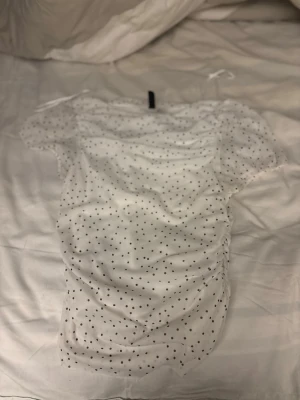 Vit prickig mesh topp med puffärm - Söt vit mesh topp med svarta prickar och puffiga korta ärmar. Toppen har rynkade sidor och tunna axelband som knyts. Perfekt för dig som gillar trendiga detaljer och en luftig känsla. Materialet är troligtvis polyester.