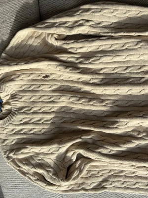 Beige kabelstickad tröja Polo Ralph Lauren - Kabelstickad beige tröja från Polo Ralph Lauren med rund halsringning och diskret broderad logga på bröstet. Tillverkad i mjukt material och har ribbade muddar vid ärmslut och nederkant. Perfekt för lager på lager och klassisk stil.
