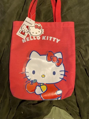 Hello Kitty röd shoppingväska - Söt röd shoppingväska med stort Hello Kitty-tryck på framsidan där Hello Kitty håller ett äpple. Väskan har två handtag och är gjord i slitstarkt tygmaterial. Perfekt för dig som vill ha en färgglad accessoar med ikoniskt motiv. Helt ny