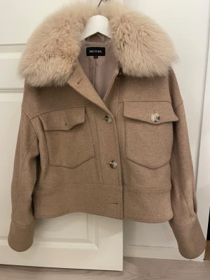 Beige ullkappa med fuskpäls från Meotine - Snygg beige ullkappa från Meotine med stor fluffig fuskpäls-krage. Kappan har två stora bröstfickor med lock, knäppning framtill och en rak, boxig passform. Perfekt för dig som vill ha en trendig och varm look i höst och vinter.