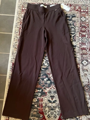 Bruna vida byxor från Cecil - Mörkbruna byxor från Cecil med vida ben och hög midja. Byxorna har en enkel design utan synliga fickor framtill och är tillverkade i ett mjukt, stretchigt material som ger en bekväm passform. Perfekta för en avslappnad och trendig look.