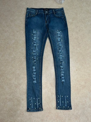 Blå  jeans med nitar och slitningar - Jeans med slitna detaljer och pärlor som ger en unik och trendig look. Helt oanvända och i nyskick. Sitter jättefint på kroppen och passar perfekt till både vardag och fest. Storlek M/L
