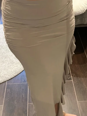Beige långkjol med volangdetalj - Snygg beige långkjol med figurnära passform och volang längs sidan. Kjolen har en lätt glansig yta och är perfekt för dig som gillar en stilren look med det lilla extra.