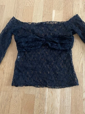 Svart spetsig offshoulder topp - Trendig svart offshoulder topp i spets med lång ärm och stor rosettdetalj framtill. Transparent spetsmaterial ger en edgy vibe och toppen är perfekt för dig som vill sticka ut. Snygg att styla med både jeans och kjol.