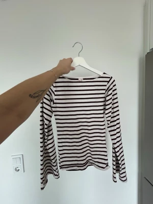 Randig långärmad topp från Nelly  - Säljer en vit långärmad topp från Nelly med mörkbruna horisontella ränder. Toppen har en klassisk, rak passform och är tillverkad i mjuk bomull. Perfekt till jeans eller kjol för en enkel och stilren look.