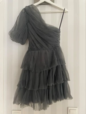 Mörkgrå one shoulder volangklänning - Snygg mörkgrå kortklänning med volanger och plisserat tyg. Klänningen har en axelbandslös design på ena sidan och ett tunt axelband på andra, vilket ger en cool one shoulder-look. Perfekt för dig som vill sticka ut med en trendig och luftig stil.