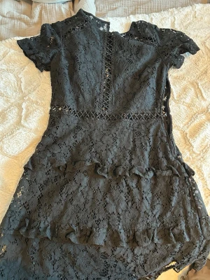 Svart spetsklänning med volang - Supersnygg svart kortklänning i spets med volangdetaljer och korta ärmar. Klänningen har fina broderade band i midjan och framtill, samt en lätt asymmetrisk nederkant. Perfekt för dig som gillar romantisk och trendig stil.