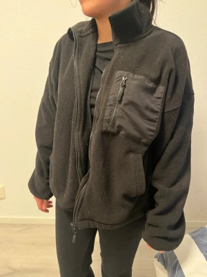 Svart fleecejacka med dragkedja - Mysig svart fleecejacka med hög krage och dragkedja framtill. Jackan har en bröstficka med dragkedja i kontrasterande material och två sidofickor. Passformen är loose och jackan är perfekt för kyliga dagar.