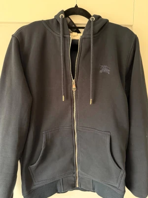Mörkblå zip hoodie från Burberry - Snygg mörkblå hoodie med dragkedja från Burberry. Klassisk modell med huva, dragsnören och två stora fickor framtill. Diskret Burberry-logga på bröstet. 