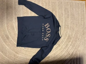 Mörkblå sweatshirt från Hugo Boss - Snygg mörkblå sweatshirt från Hugo Boss med stort BOSS-tryck på bröstet och vertikal logga. Tröjan har rund hals och långa ärmar, tillverkad i mjukt material som känns skönt mot huden. Perfekt för dig som gillar stilrena och trendiga plagg.
