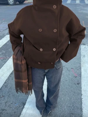 Mörkbrun dubbelknäppt rock - Stilren mörkbrun rock med dubbelknäppning och stora knappar. Rocken har en boxig passform och hög krage. Tillverkad i ett tjockare tyg som passar bra under lite kyligare dagar. Perfekt för dig som gillar en clean och modern look. 