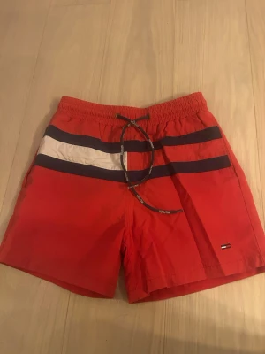 Röda badbyxor Tommy Hilfiger - Snygga röda badbyxor från Tommy Hilfiger med två mörkblå ränder och en vit rand framtill. Klassisk logga broderad på benet och svart dragsko i midjan. Perfekta för stranden eller poolen, tillverkade i snabbtorkande material.