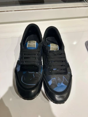 Valentino rock runners - Säljer ett par snygga Valentino rockrunners sneakers i svart med blått abstrakt mönster. Skorna har svarta skosnören, låg profil och är tillverkade i en mix av läder och textil. Perfekta för dig som vill ha något unikt och trendigt på fötterna.
