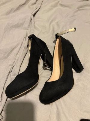 Svarta klackskor - Stilrena svarta klackskor med rund tå, bred klack och justerbar ankelrem med spänne. Skorna har en mjuk mockaliknande yta och beige innersula. Perfekta för dig som vill ha både höjd och stabilitet i steget.