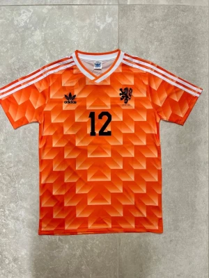 Holland retro matchtröja Adidas - Säljer en ikonisk orange Holland retro matchtröja från Adidas med nummer 12 och namnet Van Basten på ryggen. Tröjan har ett geometriskt mönster, vita ränder på axlarna och svart tryck. Perfekt för fotbollsfans som gillar klassiska landslagströjor.  Finns i olika storlekar!!! Pris kan diskuteras!!!
