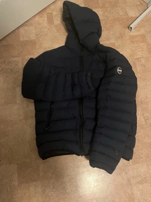 Marinblå  pufferjacka från Colmar - Snygg marinblå pufferjacka från Colmar med huva och dragkedja framtill. Jackan har ett broderat Colmar-märke på ärmen och två praktiska fickor med dragkedja. Perfekt för kalla dagar när du vill hålla värmen och ändå se stilren ut.