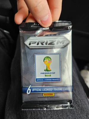Panini Prizm VM 2014 samlarkort pack SEALED - Oöppnat paket med 6 officiella samlarkort från Panini Prizm, FIFA World Cup Brasil 2014. Förpackningen är silverfärgad med VM-logga och Panini-märke. Perfekt för dig som gillar fotboll och samlarprylar.