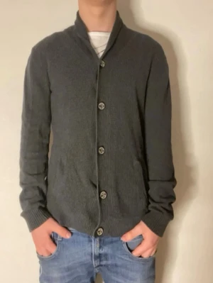 Mörkgrå kofta från Tom Tailor - Mörkgrå kofta från Tom Tailor med knäppning framtill och ribbade muddar. Klassisk och enkel design med smal passform och mjukt material. Perfekt att slänga över en t-shirt för en avslappnad look.