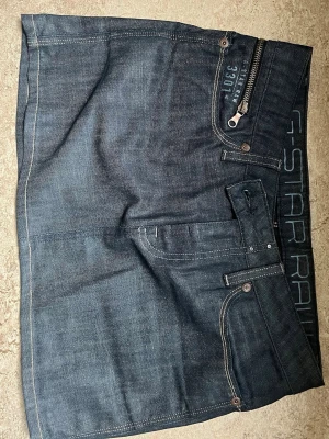 Mörkblå jeanskjol från G-Star RAW - Snygg mörkblå jeanskjol från G-Star RAW med klassiska fickor fram och bak, dekorativa sömmar och detaljer som dragkedja och knappar. Kjolen har en kort modell och är perfekt för dig som gillar streetwear och denim. Dragkedjan går att fixa den jag hoppat ur och jag pallade inte sätta tillbaka den 