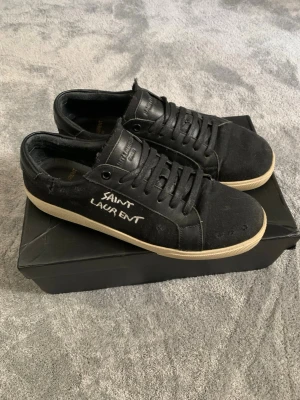 Svarta Saint Laurent sneakers - Svarta sneakers från Saint Laurent med vit text på sidan. Skorna har en klassisk låg siluett, svart canvas och läderdetaljer samt vita/beige skosulor. Snygg och stilren design med snörning framtill och diskret logga på plösen. Hör av dig vid minsta fundering och pris kan diskuteras! 🤩💯