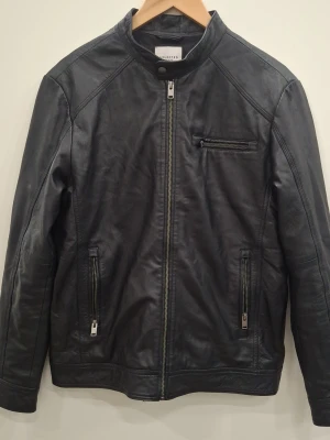 Svart skinnjacka från Selected Homme - Snygg svart skinnjacka från Selected Homme med rak passform, ståkrage och dragkedja framtill. Jackan har tre fickor med dragkedja och diskreta sömdetaljer för en stilren look. Perfekt för dig som vill ha en klassisk bikerstil.